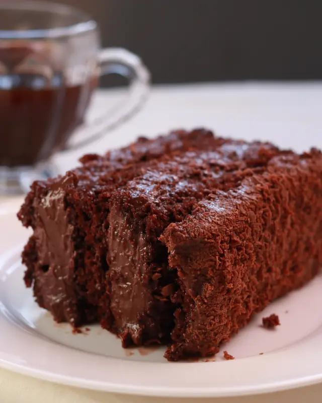 Fatia de bolo de chocolate num prato branco sobre toalha clara, com xícara de vidro ao fundo