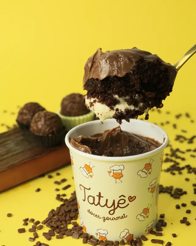 Colher erguendo uma porção de massa de chocolate com creme branco sobre o pote da Tatyê, ao lado de docinhos de chocolate